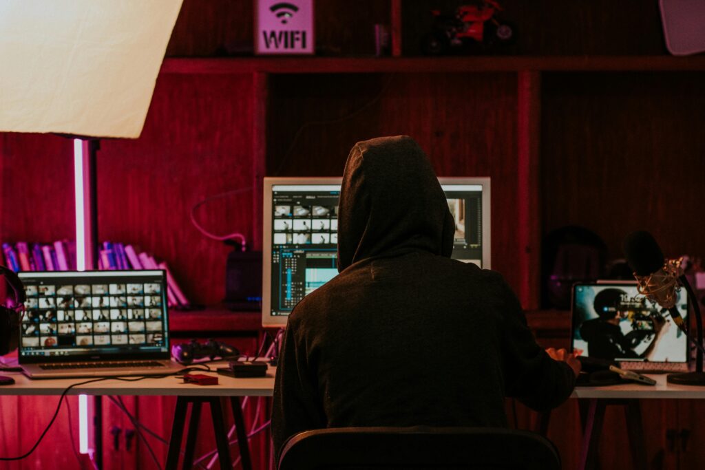 Person in a hoodie editing videos on multiple screens in a dimly lit space with neon lighting.